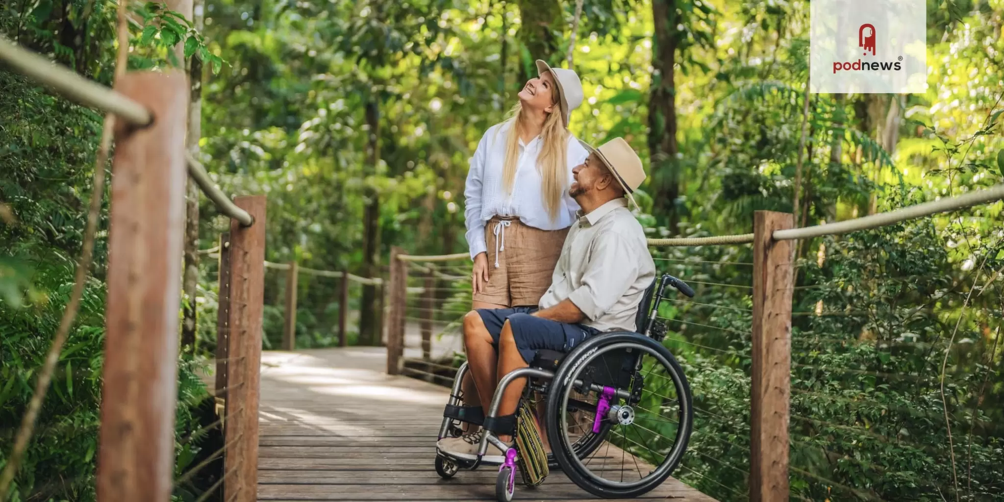 Man in wheelchair with woman on boardwalk in forested area looking up at the trees and smiling with Podnews logo superimposed on top right. 